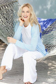 A blonde woman wearing a white top and a light blue cardigan sitting in a hammock