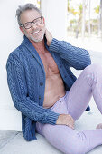 A grey-haired man with a beard and glasses wearing a cardigan and trousers