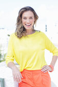 A dark-blonde woman wearing a yellow blouse and orange shorts