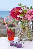 A table at the beach with a bouquet of roses, dahlia, and phlox, and a cocktail