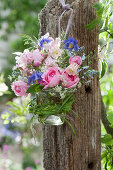 A small bouquet of roses, columbines, speedwell, and flowers of caraway in a glass jar with a grass wreath hung on a post