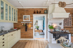 Cream-coloured kitchen units with glass-fronted wall units, rough wooden wall and iron stove in open-plan kitchen