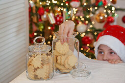 A boy wearing a Santa hat reaching into a jar of biscuits