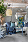 Old sofa with decorative cushions under round mirror in open living room with concrete floors and wooden beams