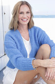 Langhaarige Frau in weißem Sommerkleid und blauer Strickjacke am Strand sitzend