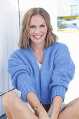 Langhaarige Frau in weißem Sommerkleid und blauer Strickjacke am Strand sitzend