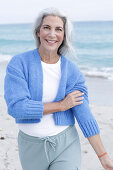 Reife Frau mit grauen Haaren in weißem T-Shirt, blauer Strickjacke und Hose am Strand