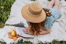 Female in dress with straw hat on face lying with open book on picnic blanket while chilling alone and enjoying summer weekend in countryside