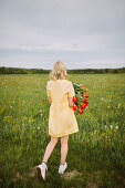 Back view female in dress standing with bunch of red tulip flowers in meadow in summer and looking away