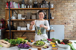 Happy female mixing tasty vegetable salad with lettuce leaves at table in loft style house