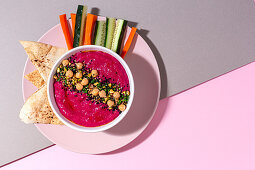 Beetroot hummus garnished with chickpea served on two colored background with bread and fresh carrot and cucumber sticks