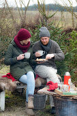 Mann und Frau mit Glühwein und Weihnachtsgebäck auf Bank sitzend im Garten