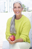 Grauhaarige Frau mit einem Apfel in grüngelbem Strickpullover und heller Hose