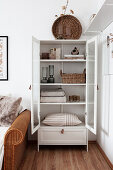 White cabinet with glass doors holding books, decorative objects, table linen, and cushions next to rattan sofa