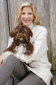 A mature blonde woman holding a dog wearing a white knitted jumper and grey leggings