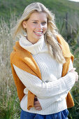 Young blond woman in white turtleneck sweater, with yellow knitted sweater slung over shoulders in nature