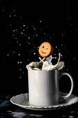 Smiling and winking cookie dropping in ceramic mug of milk with splatters in motion on black background