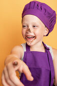 Laughing boy in purple chef uniform with face covered with chocolate pointing at camera and looking away against yellow background