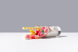 Fallen glass with bunch of sweet gummy candies and white sugar placed near striped straw on grey and white background