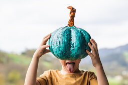 Unrecognizable funny kid showing tongue while covering face with teal pumpkin in raised arms in rural area on blurred background