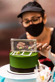 Konditorin mit Gesichtsmaske beim Verzieren einer Torte mit Tennisplatz-Motiv