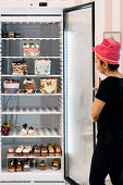 Hispanic woman in headscarf opening refrigerator and checking creative cakes and cupcakes during work in bakery