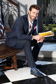 Young man with a beard in a jacket and jeans sitting on a leather sofa with a book in his hands