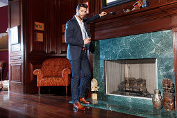 Young man with a beard in a jacket and jeans is standing in front of a marble fireplace