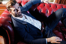 Young man with a beard in a jacket and jeans lies on a leather sofa