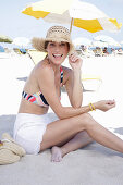 Junge brünette Frau mit Sommerhut in gestreiftem Bikinioberteil und weißen Shorts am Strand
