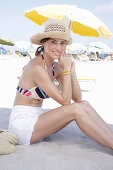 Junge brünette Frau mit Sommerhut in gestreiftem Bikinioberteil und weißen Shorts am Strand