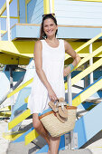 Brünette Frau in weißem Sommerkleid mit Flipflops und Korbtasche am Strand