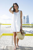 Brünette Frau in weißem Sommerkleid mit Flipflops und Korbtasche am Strand