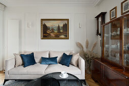 Helles Sofa mit Kissen vor antiker Anrichte, Gemälde an weißer Wand