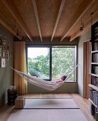 Narrow room with wood ceiling and a person lying in a hammock in front of window with garden view