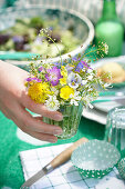 Hand holding spring bouquet of wild flowers with dandelion, cranesbill and horned violet in a small glass