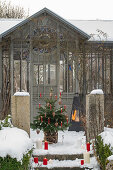 Geschmückter Christbaum mit Wachskerzen auf verschneiter Terrasse und Winterfeuer