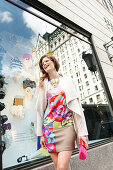 Young woman with glasses in a colorful dress and a light coat in front of a shop window