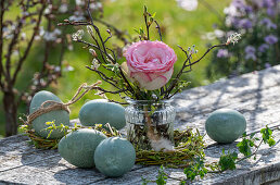 Rosenblüte (Rosa) mit Zweigen in Vase und Ostereiern auf Gartentisch