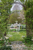 Gedeckter Tisch im Garten für Osterfrühstück mit Osternest und gefärbten Eiern, Blumenstrauß, Korb mit Eiern in der Wiese, Torbogen durch Kletterpflanzen