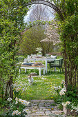Gedeckter Tisch im Garten für Osterfrühstück mit Osternest und gefärbten Eiern, Blumenstrauß, Hund in der Wiese, Torbogen durch Kletterpflanzen