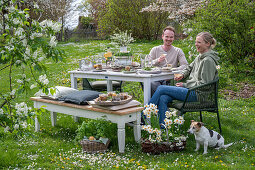 Junges Paar sitzt an gedecktem Tisch zum Osterfrühstück mit Osternest und gefärbten Eiern in Eierbechern, Sektgläser, Narzissen und Petersilie im Korb, mit Hund im Garten