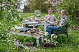 Young couple sitting at a laid table for Easter breakfast with Easter nest and colored eggs, toasting with champagne, daffodils and parsley in a basket, with dog in the garden