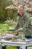 Junge Frau baut Etagere aus Blumenstrauß auf Suppenschüssel, gedeckter Tisch zum Osterfrühstück mit Osternest und gefärbten Eiern und Wasserkrug