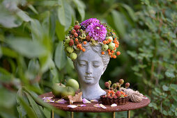 Bust of a woman decorated with berries, ornamental apples and dahlia on garden table