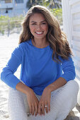 Woman in blue jumper and white trousers sitting on beach