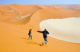 Eine junge Frau und ein junger Mann rennen über die steile Flanke des Big Daddy hinunter. Eine der höchsten Dünen der Erde. Tief unten die Ebene Deadvlei. Die Sossusvlei Dünen. Namib Wüste. Namibia. Afrika. MR