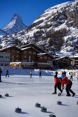 People curling on a rink, Matterhorn in background, Zermatt, Valais, Switzerland (Curling: A rink game where round stones are propelled by hand on ice towards a tee (target) in the middle of a house (circle)).