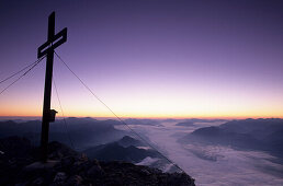 The break of dawn at a cross on the summit of Grimming with view over a fogbank into the Enns valley towards Gesaeuse, Dachsteingruppe, Styria, Austria