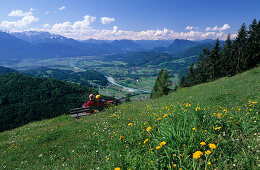 Aussichtsbank mit zwei Wanderer in eine Blumenwiese am Kranzhorn und Blick auf Inntal, Kaisergebirge und Zentralalpen, Chiemgauer Alpen, Tirol, Österreich